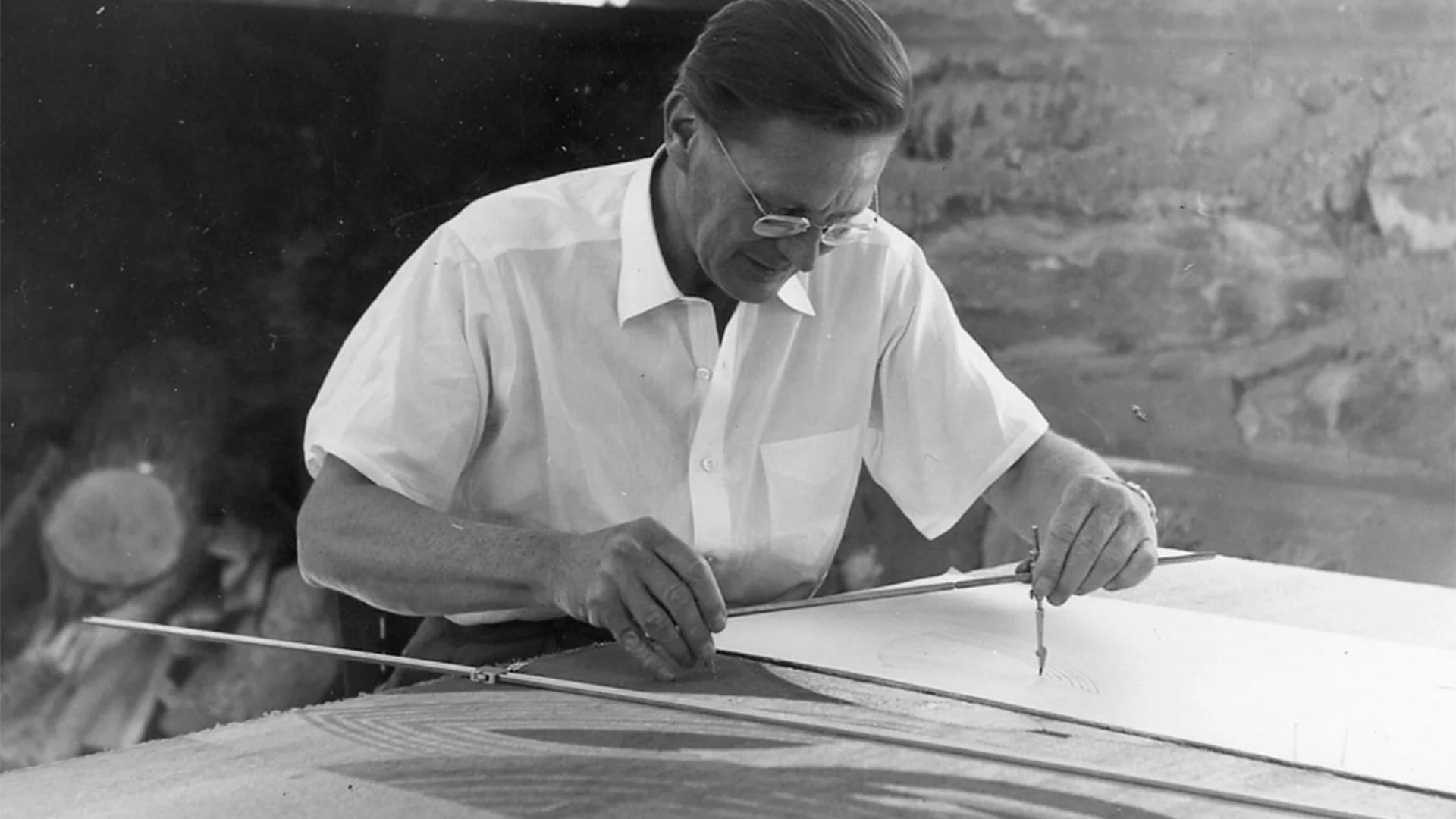 A person uses a compass to draw on paper, focusing intently. They wear glasses and a light shirt, sitting at a table against a rustic background.