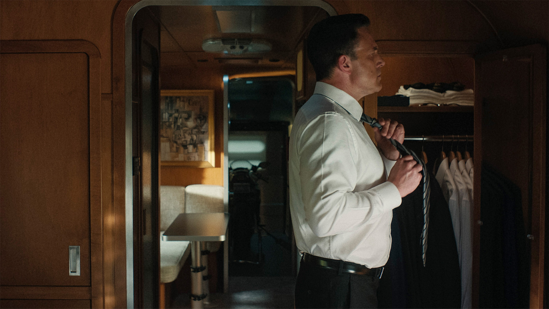 Man adjusting his tie in a dimly lit room with wooden walls and a closet full of shirts.