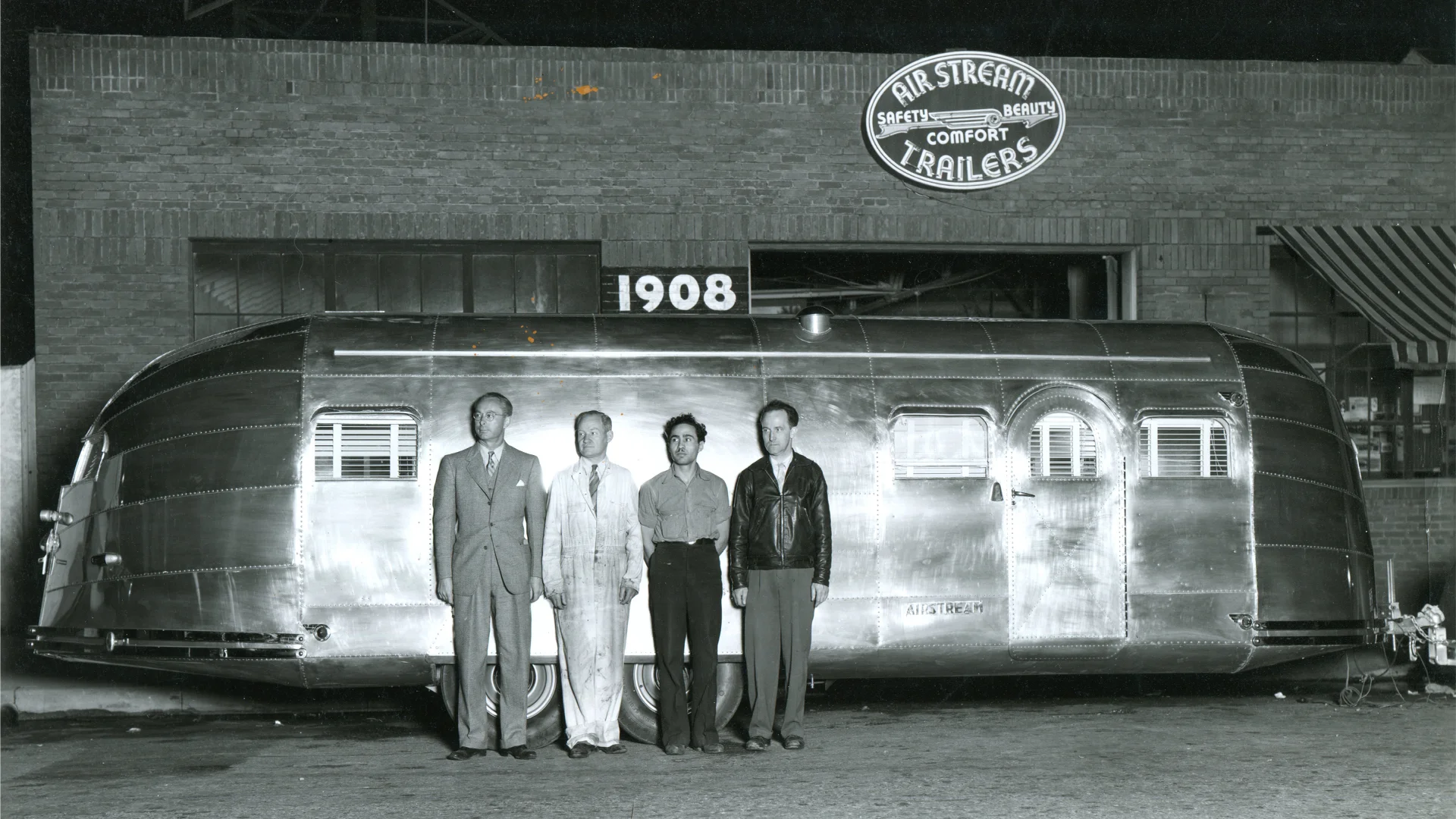 Four men stand in front of a shiny, vintage Airstream trailer parked outside a brick building with a sign reading Airstream Trailers and a large 1908 above the trailer. The image is black and white.