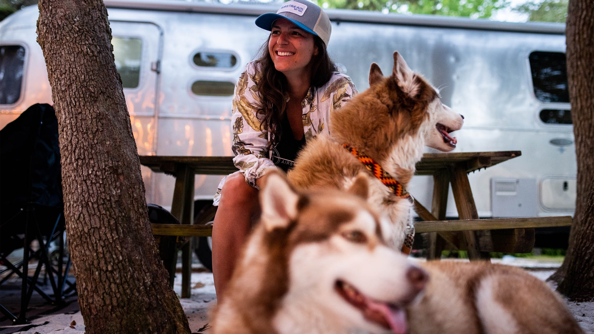 A woman wearing a hat and patterned shirt sits at a picnic table outdoors, smiling beside two brown and white huskies, with an Airstream trailer in the background.