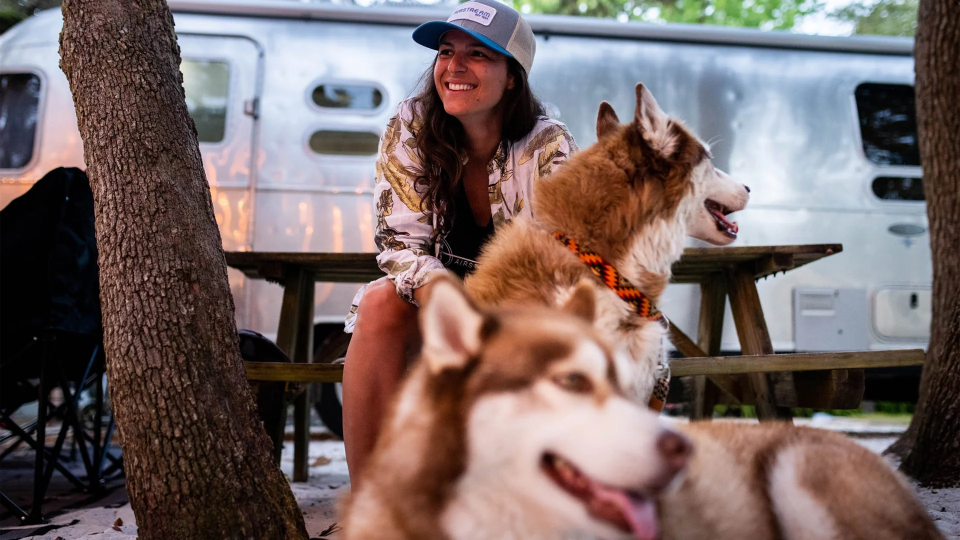 Woman in a cap sits smiling between two huskies, with a silver Airstream trailer in the background. Trees are around them.
