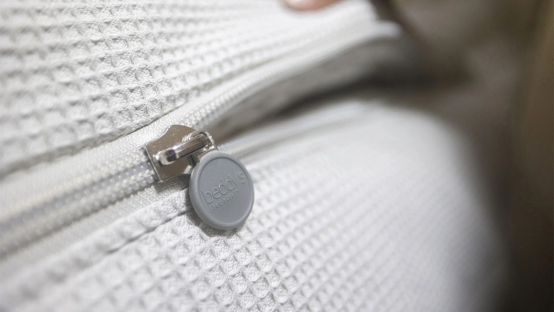 Close-up of a gray zipper with a round pull tag on a textured fabric cover. The tag displays the word "bedgear."