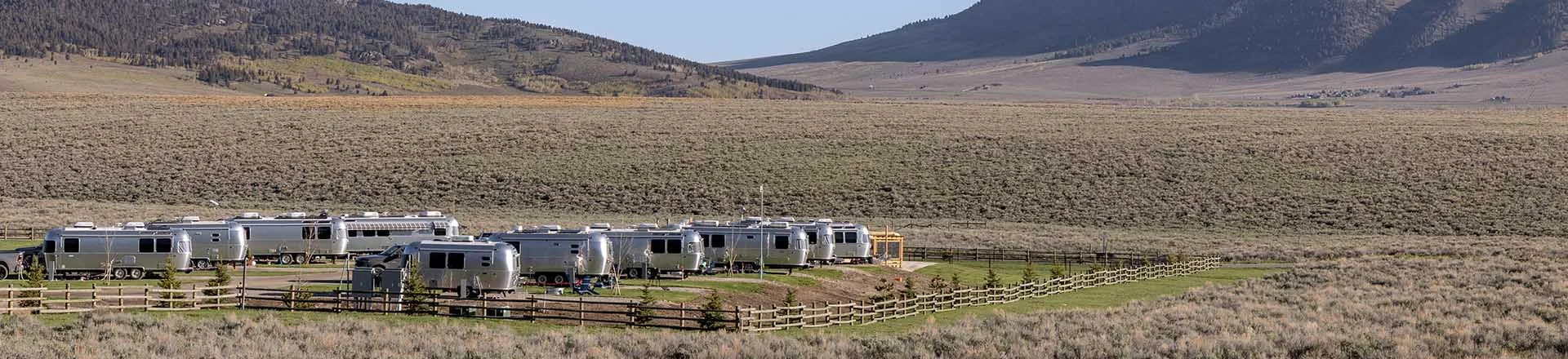 Several silver Airstream trailers are parked in a fenced area on a grassy field, surrounded by sagebrush and mountains under a clear blue sky.