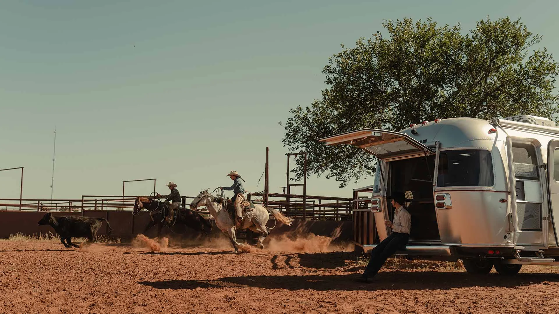 Two cowboys on horseback chase a bull in a dusty arena, with a person seated by a trailer under a clear blue sky.