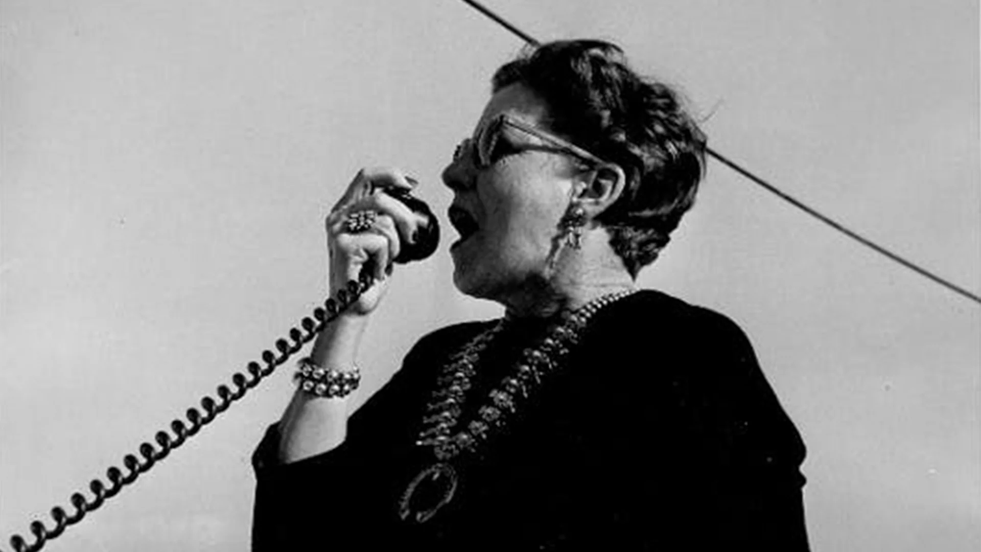 A woman with sunglasses and jewelry speaks passionately into a microphone against a clear sky.