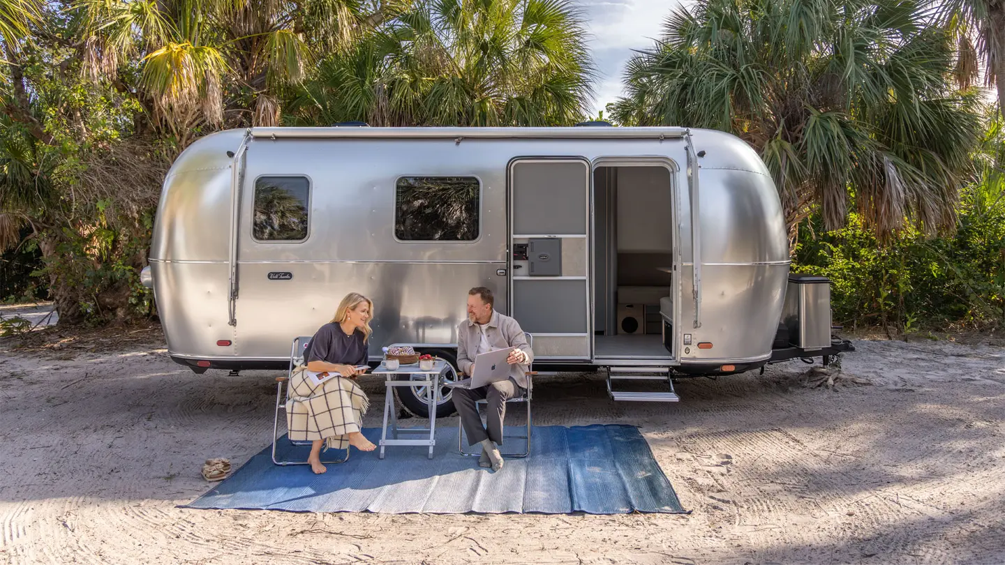A couple seated on folding chairs outside a silver Airstream trailer, surrounded by palm trees, enjoying a drink and conversation.