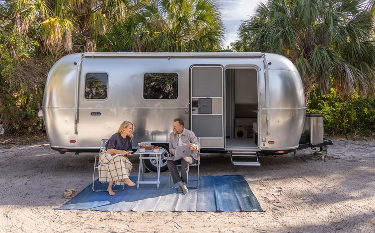 A man and woman sit on chairs outside a silver Airstream trailer, talking and using a laptop on a small table, surrounded by palm trees on a sandy ground with a blue rug beneath them.