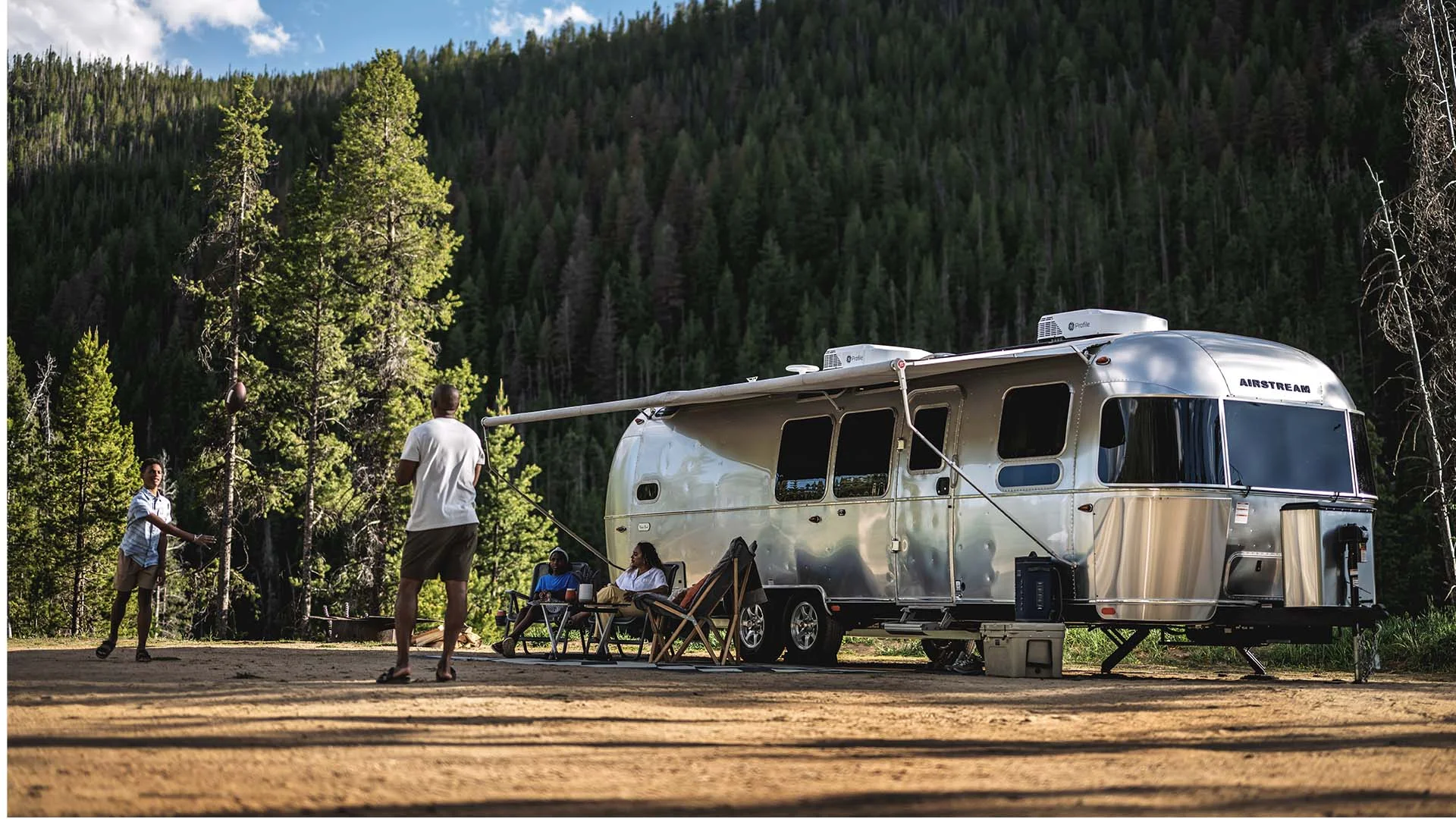 A silver Airstream trailer parked in a forested area with people sitting outside and some standing, surrounded by tall green trees.