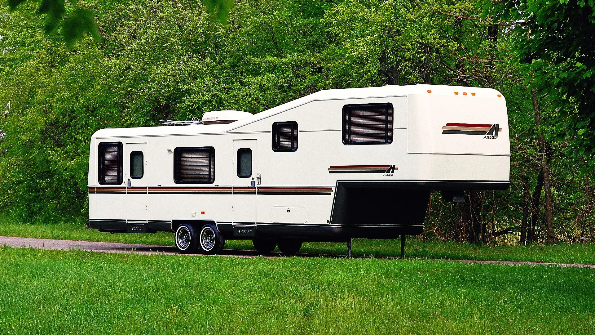 A large white fifth-wheel travel trailer with black and brown stripes is parked on a grassy area beside a paved road, surrounded by green trees and foliage.