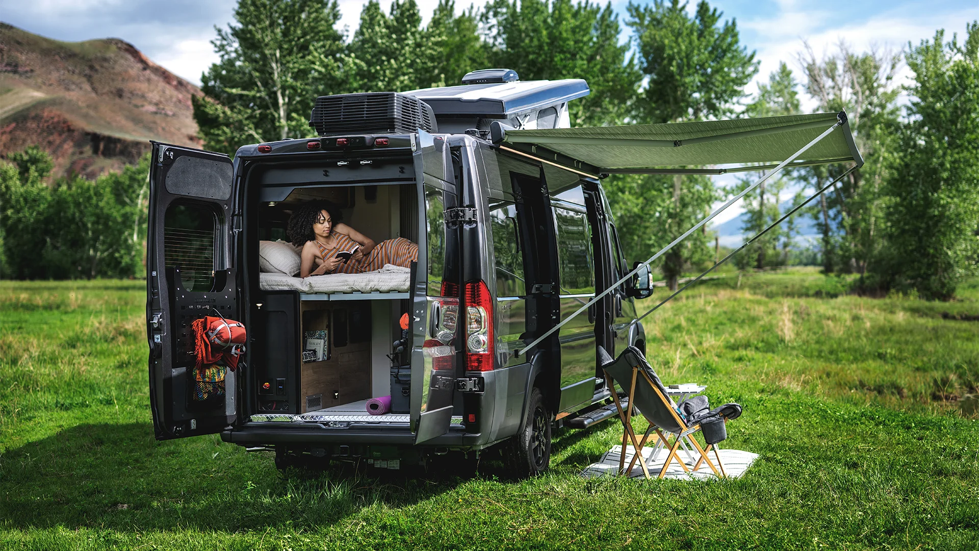 Camper van parked on grassy field, awning extended, with a person relaxing inside. Two chairs and a small table set up outside under trees.