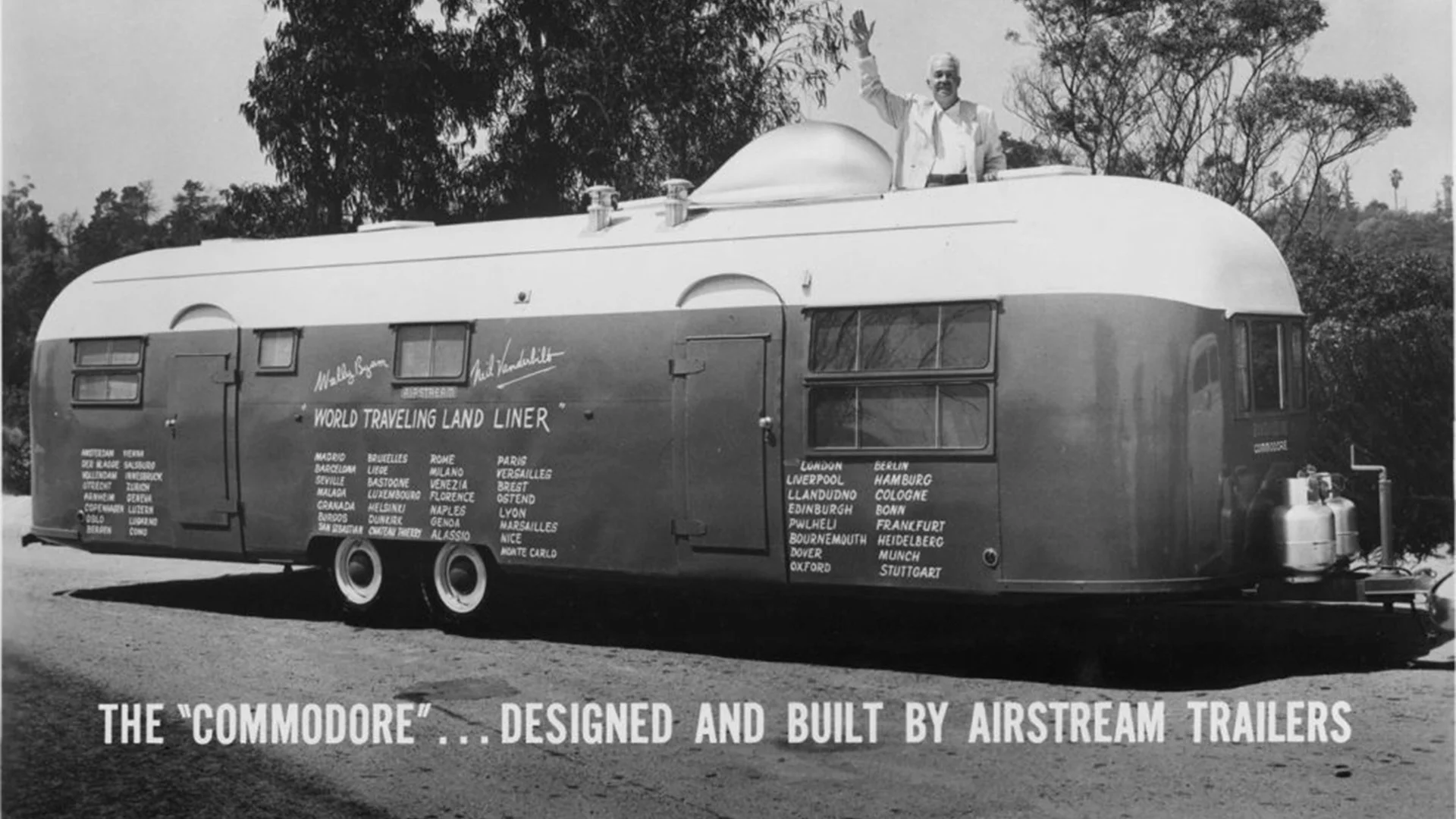 Black and white photo of a vintage Airstream trailer named "The Commodore," with a man standing on top, surrounded by trees.
