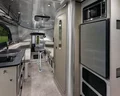 Interior of a modern travel trailer with a kitchenette, microwave, fridge, and dining area. Sleek finishes, large windows, and seating offer a spacious, well-lit, and comfortable space for travel or camping.