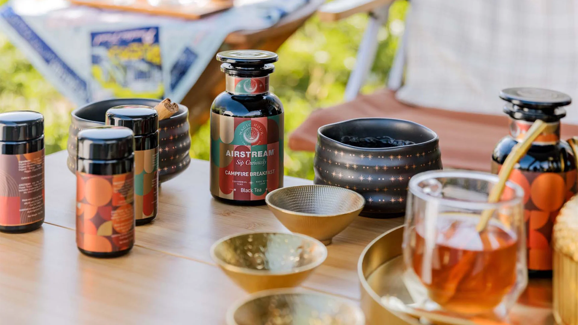 Various tea bottles and cups are arranged on a table outdoors, with a glass of tea and decorative bowls nearby.