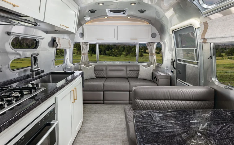 Modern RV interior with a sleek kitchen, black countertops, stainless steel appliances, a gray leather sofa, dining area, and large windows showing a scenic grassy landscape outside.