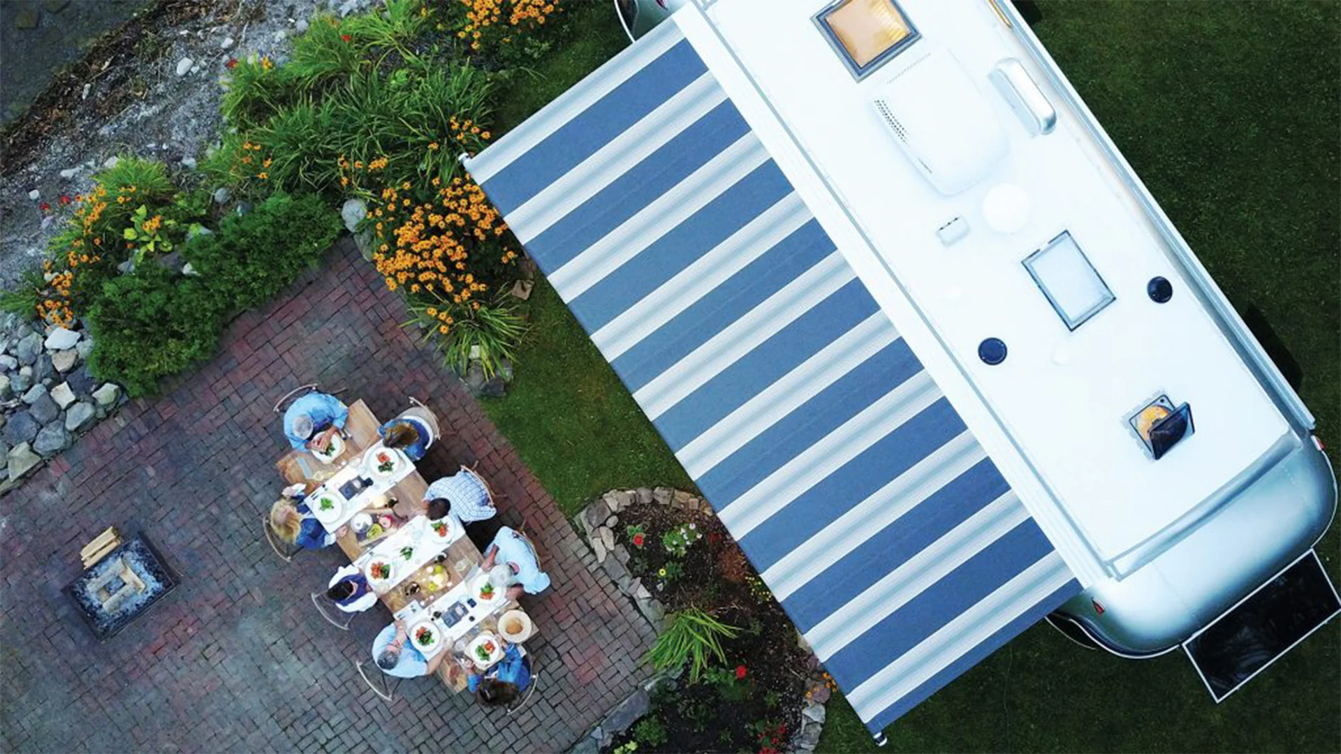 Aerial view of people dining outdoors on a brick patio next to an RV with a striped awning and surrounded by lush greenery and flowers.