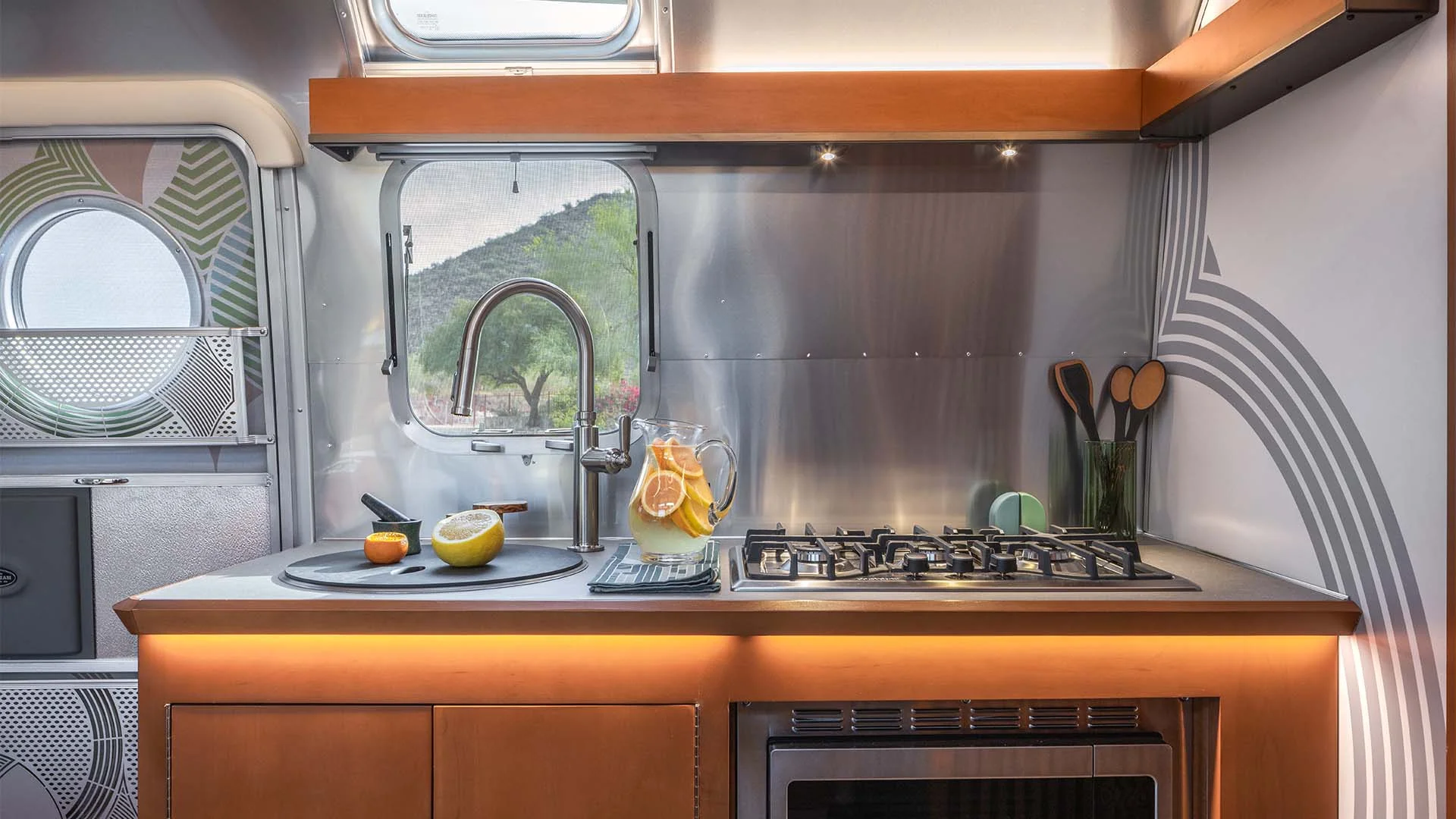 Modern RV kitchen with a gas stove, fruit, pitcher, cutting board, and scenic outdoor view through a circular window.
