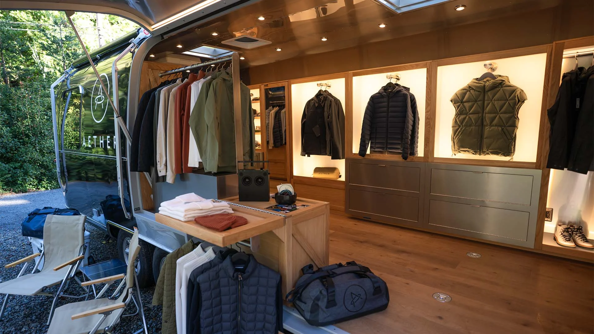 A mobile retail shop features neatly displayed jackets, shirts, and outerwear on shelves and hangers, with folded clothes on a central wooden table and duffel bags nearby. Camping chairs are set up outside on a gravel surface.