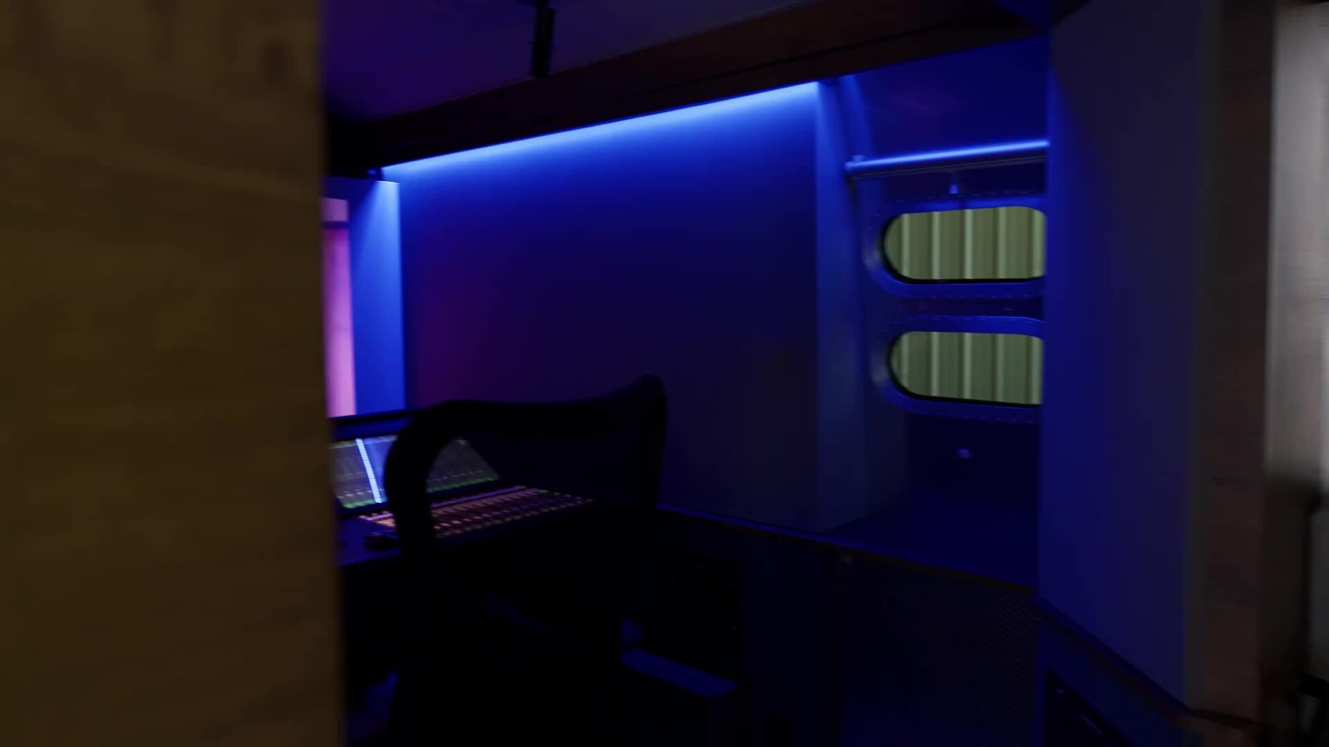 Dimly lit room with blue ambient lighting, showing a silhouetted chair and desk with computer screens, and two small windows on the wall.
