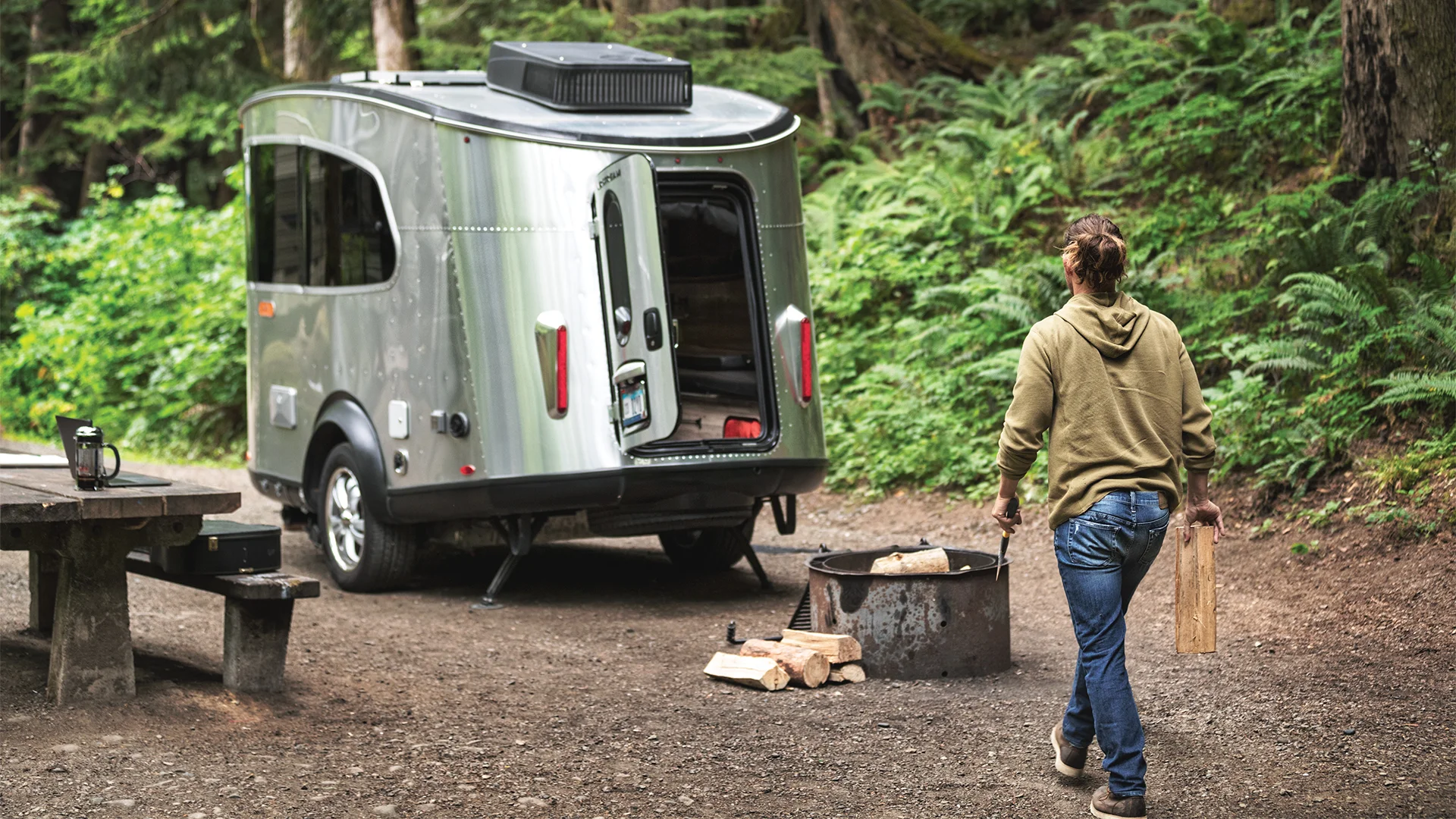 A person carries firewood toward a sleek, modern trailer parked at a wooded campsite with a picnic table and fire pit.