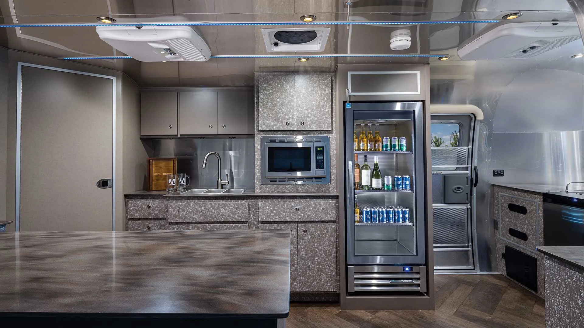 The interior of an Airstream Commercial custom bar and beverage trailer with sleek countertops, built-in microwave, stocked fridge, dual sinks, and overhead storage, featuring ambient ceiling lighting.