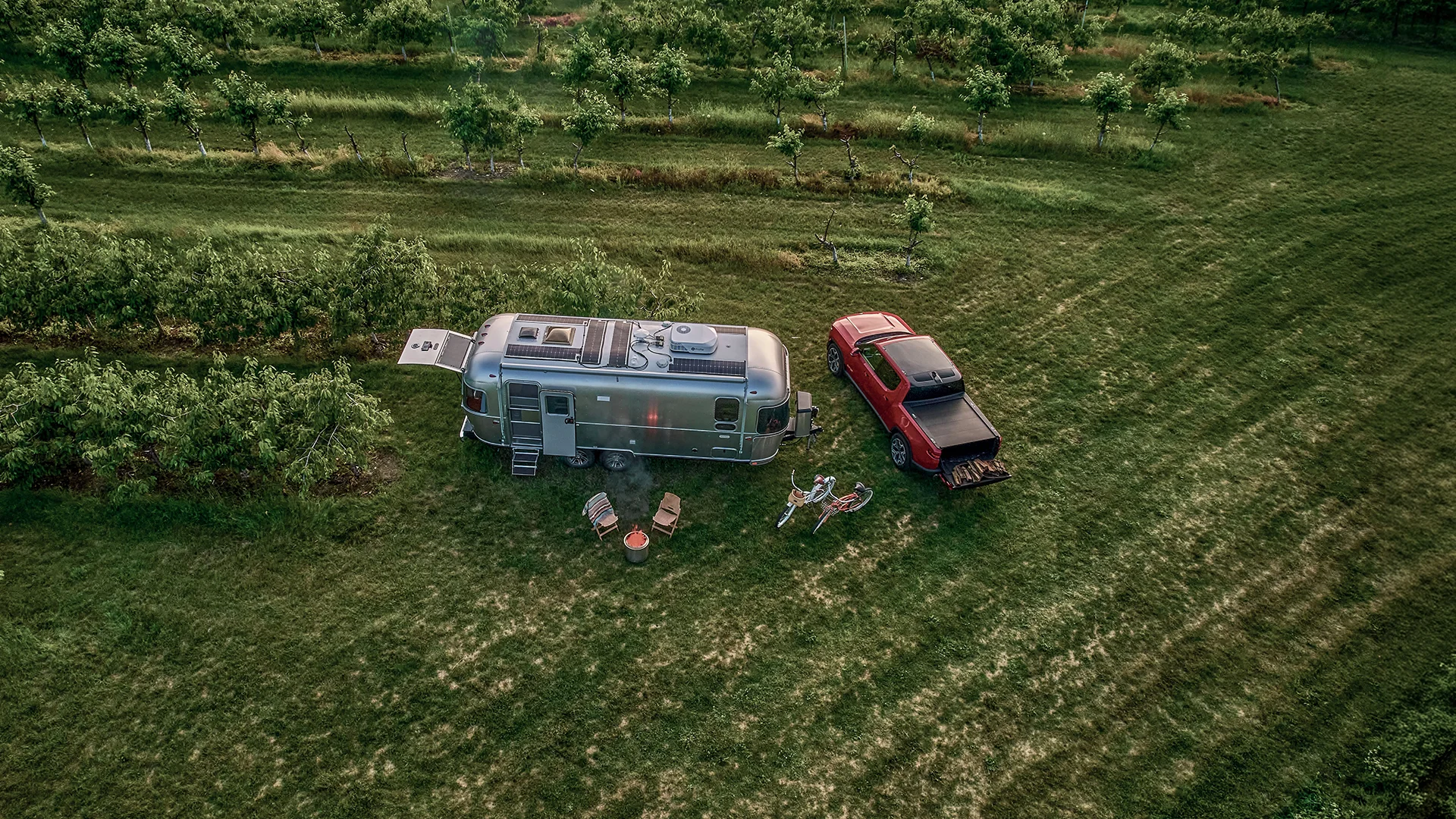 Aerial view of a silver travel trailer hitched to a red pickup truck parked on green grass, with outdoor chairs, a table, and bicycles arranged nearby, surrounded by rows of trees in a field.