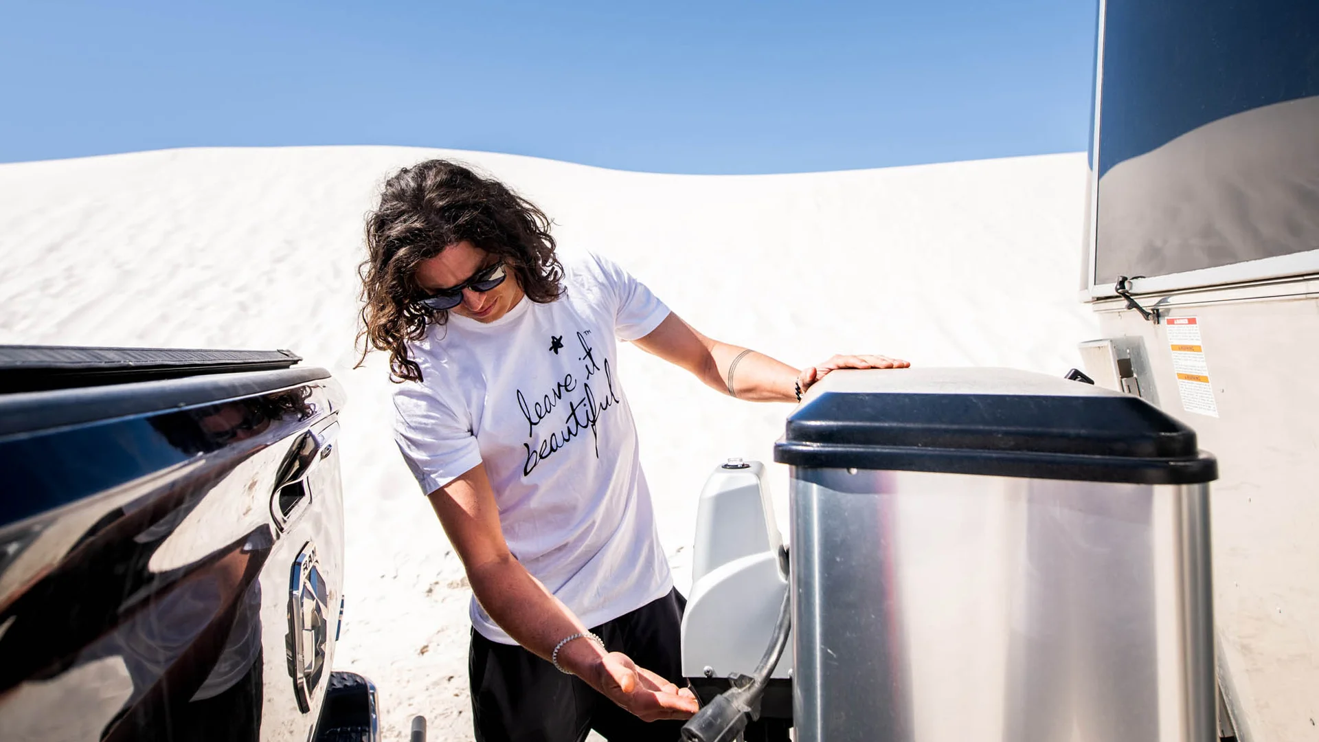 Woman with curly hair, wearing sunglasses and a "love is beautiful" T-shirt, connecting a hose to a tank in a desert area.