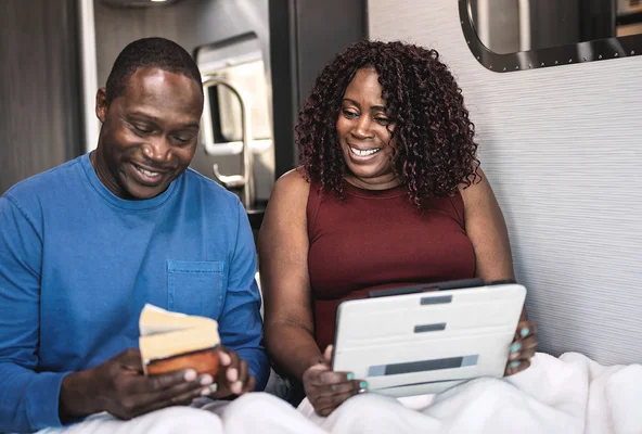 A couple sits in bed in an RV, smiling. The man holds a small book, and the woman is using a tablet.