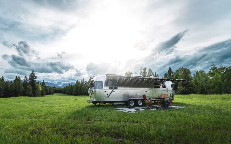 A silver RV with an awning parked on a lush green field with chairs, surrounded by trees and mountains under a cloudy sky.