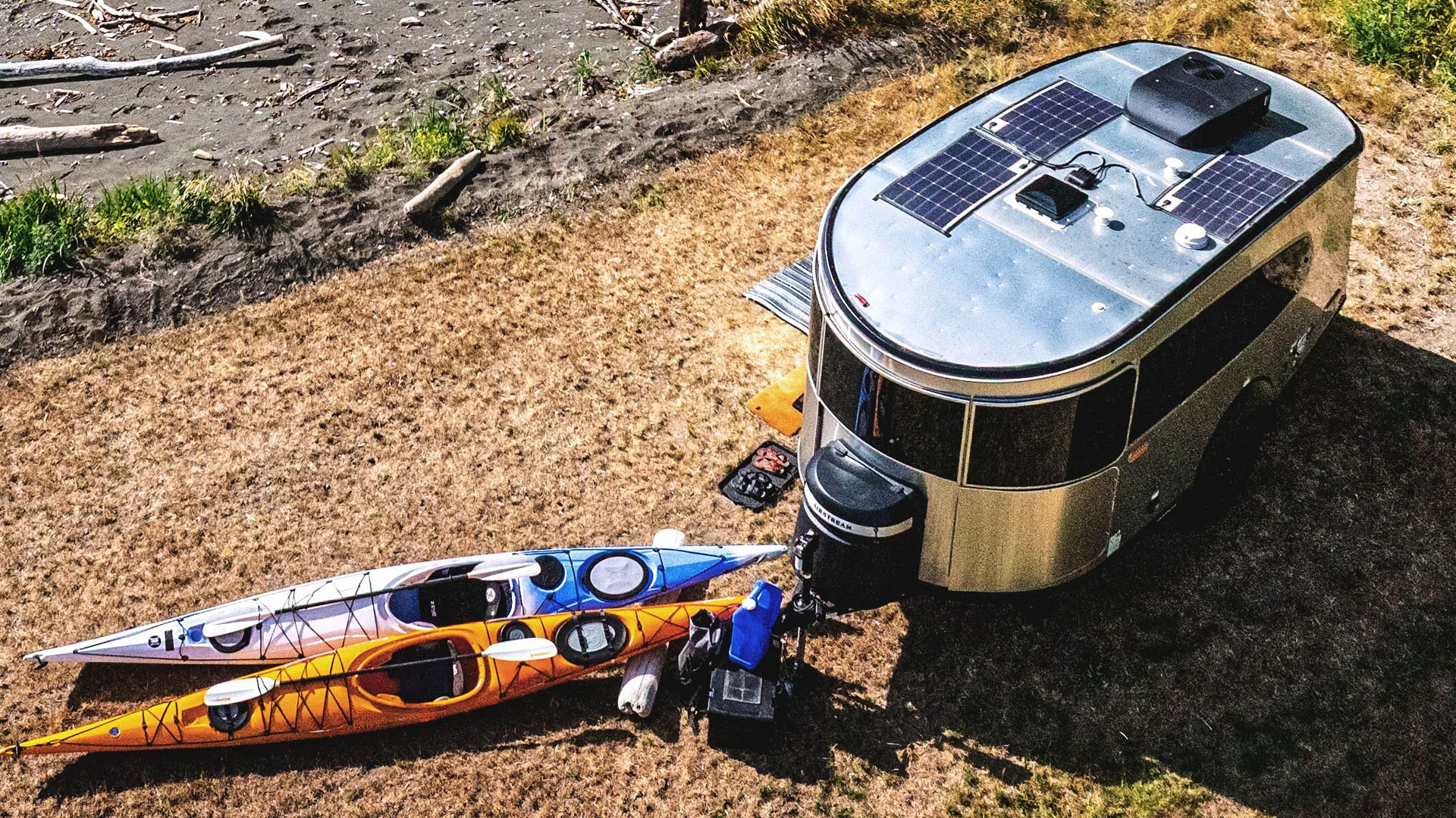 A silver camper with solar panels is parked on dry grass near a riverbank. Two colorful kayaks, one blue and one orange, are lying next to the camper, along with various outdoor gear and supplies.