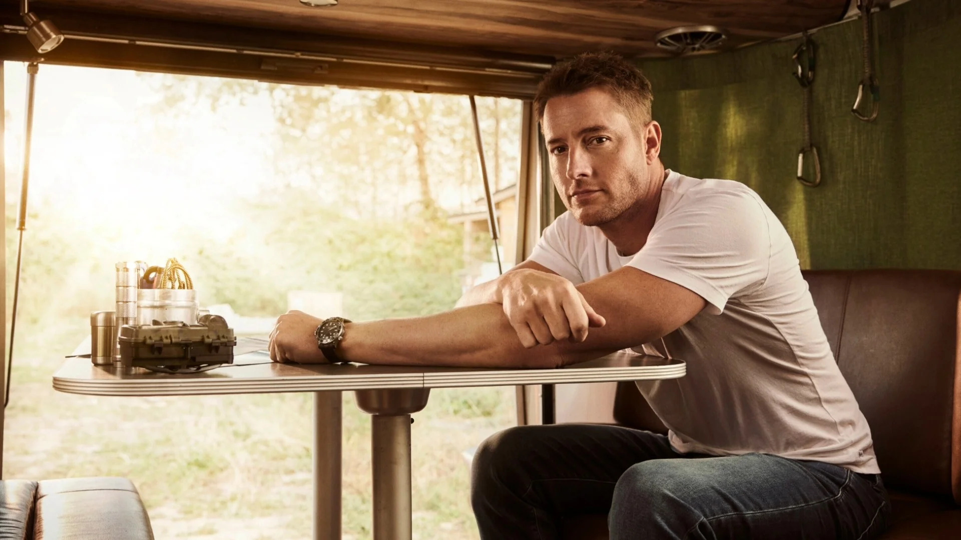 Man in a white t-shirt sits at an RV table with gadgets. Sunlight streams through the window, creating a warm ambiance.