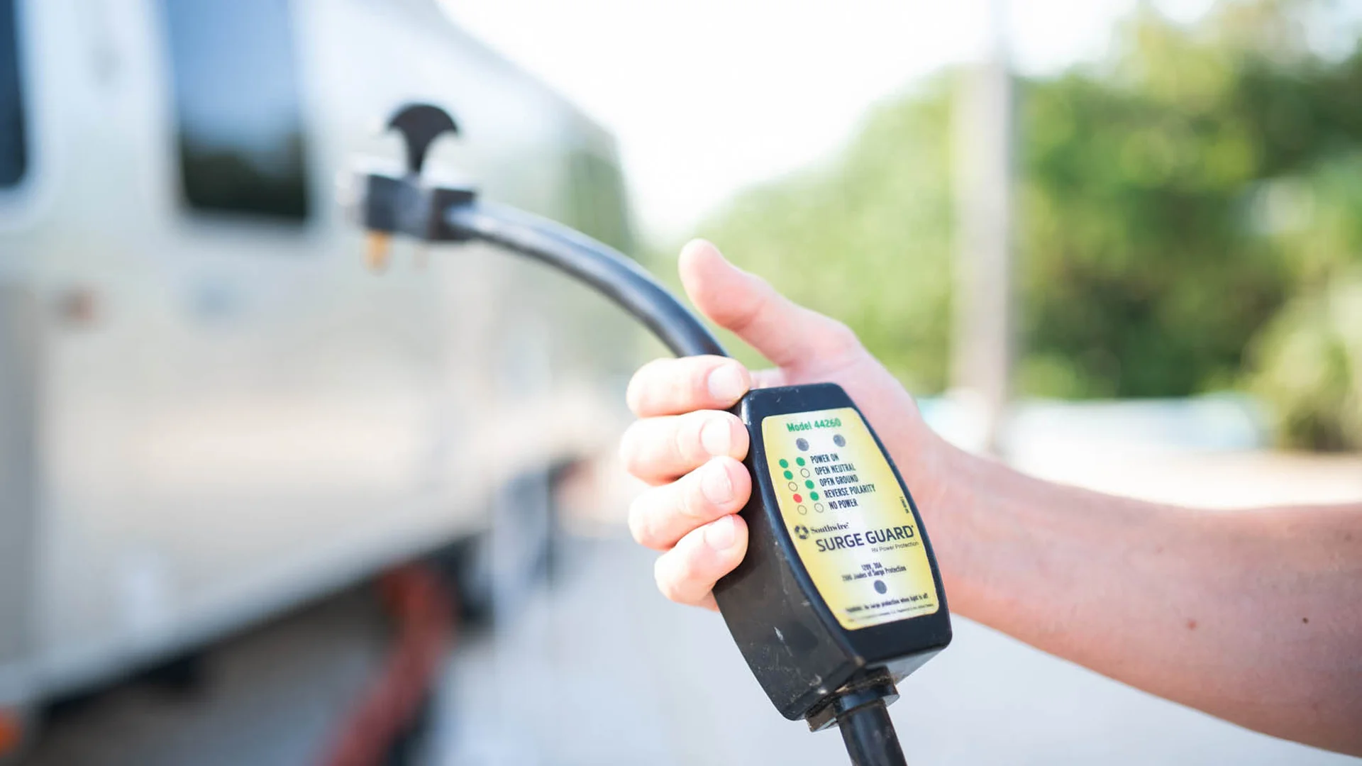 A person’s hand holds a Surge Guard electrical analyzer connected to a power cord near an RV, with trees and a bright sky in the background.