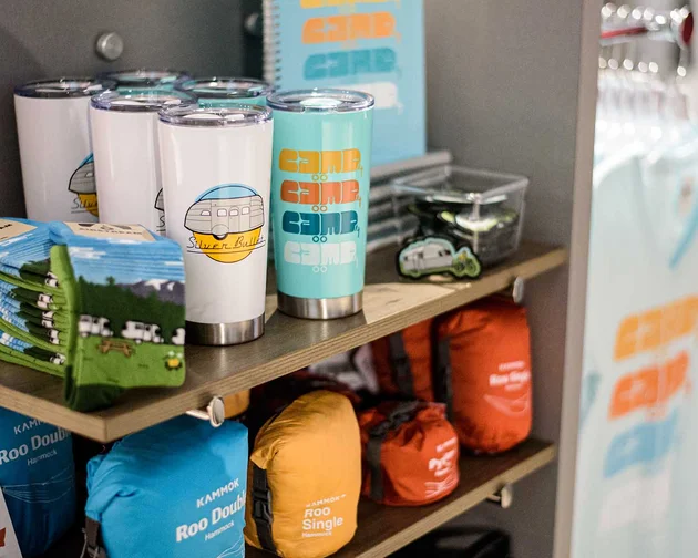 A shelf displaying tumblers with camper designs, folded t-shirts, colorful drawstring bags, and small packets in a store.