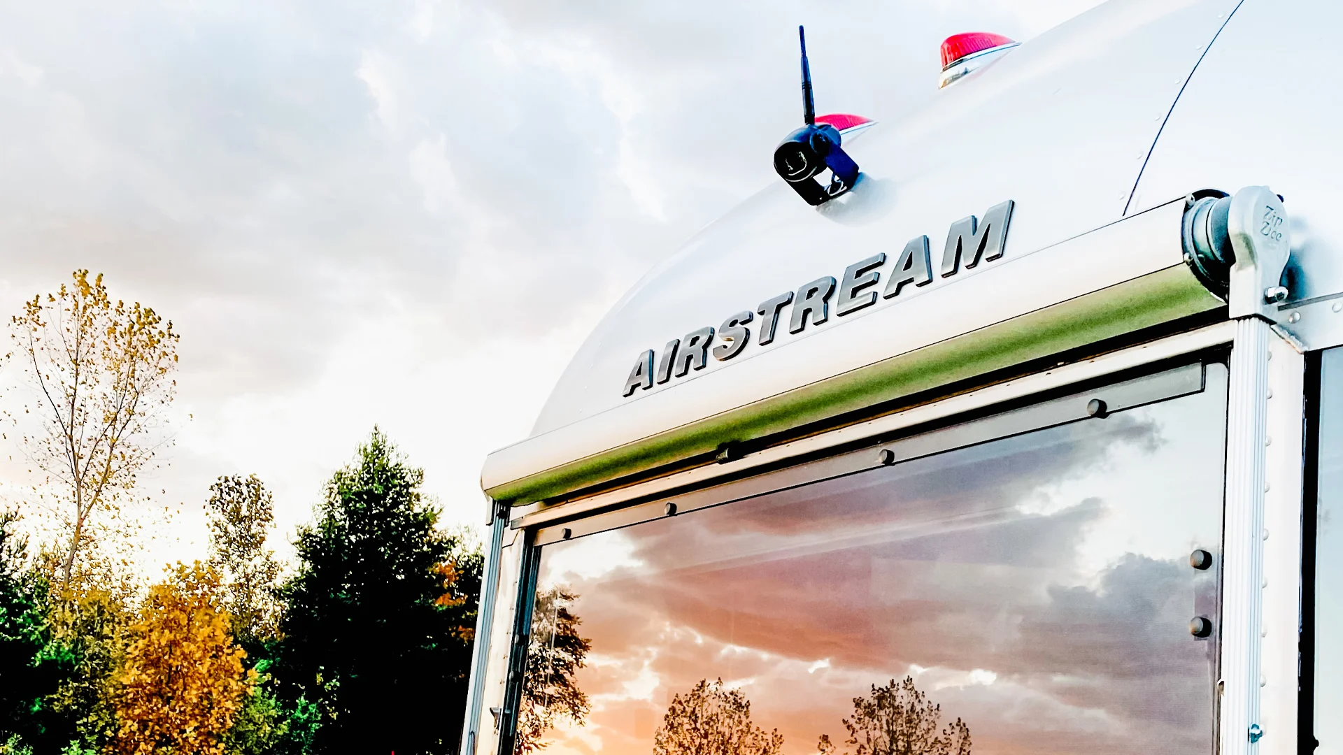 Close-up of an Airstream trailer with a reflective window showing trees and a cloudy sky at sunset. The Airstream logo is visible above the window. Trees with autumn colors appear in the background.