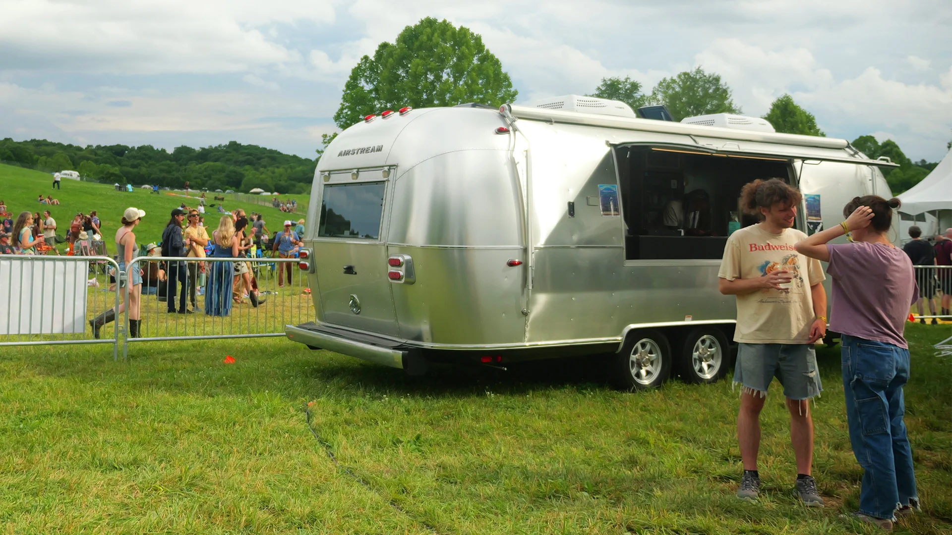 People gather around a silver Airstream Custom Bar and Beverage Trailer at an outdoor music festival, with trees and tents in the background under a cloudy sky.
