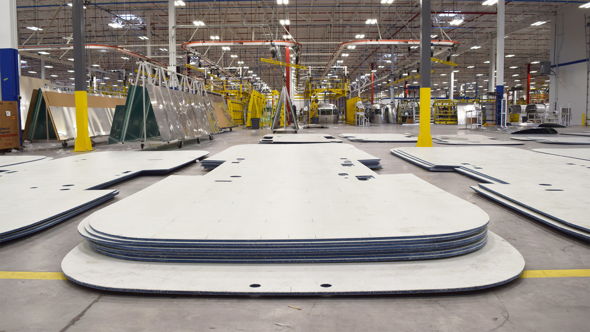 Large metal sheets stacked in a spacious, well-lit factory, surrounded by machinery and yellow safety columns.