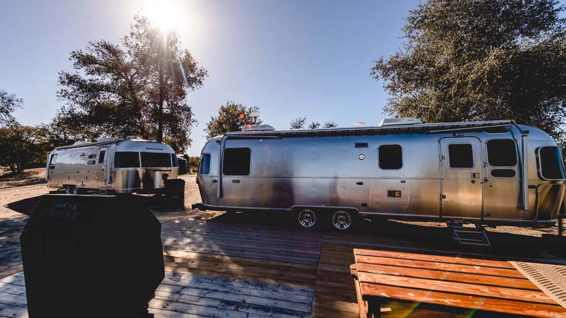 Two shiny Airstream trailers parked on a wooden deck under sunny skies, surrounded by trees. A grill cover is in the foreground.