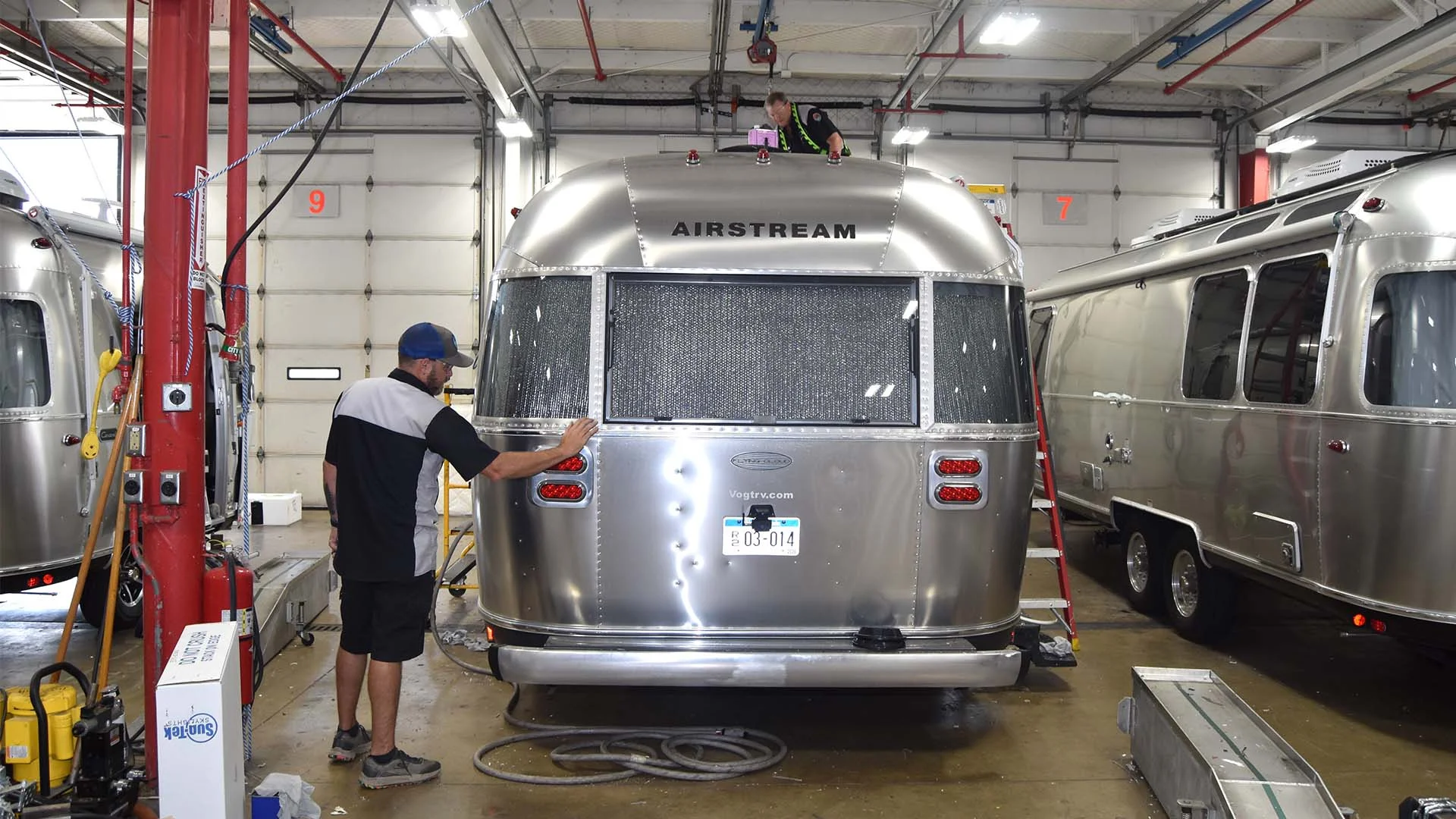 Worker inspecting a silver Airstream trailer in a repair facility, under bright ceiling lights, with tools and equipment visible around.