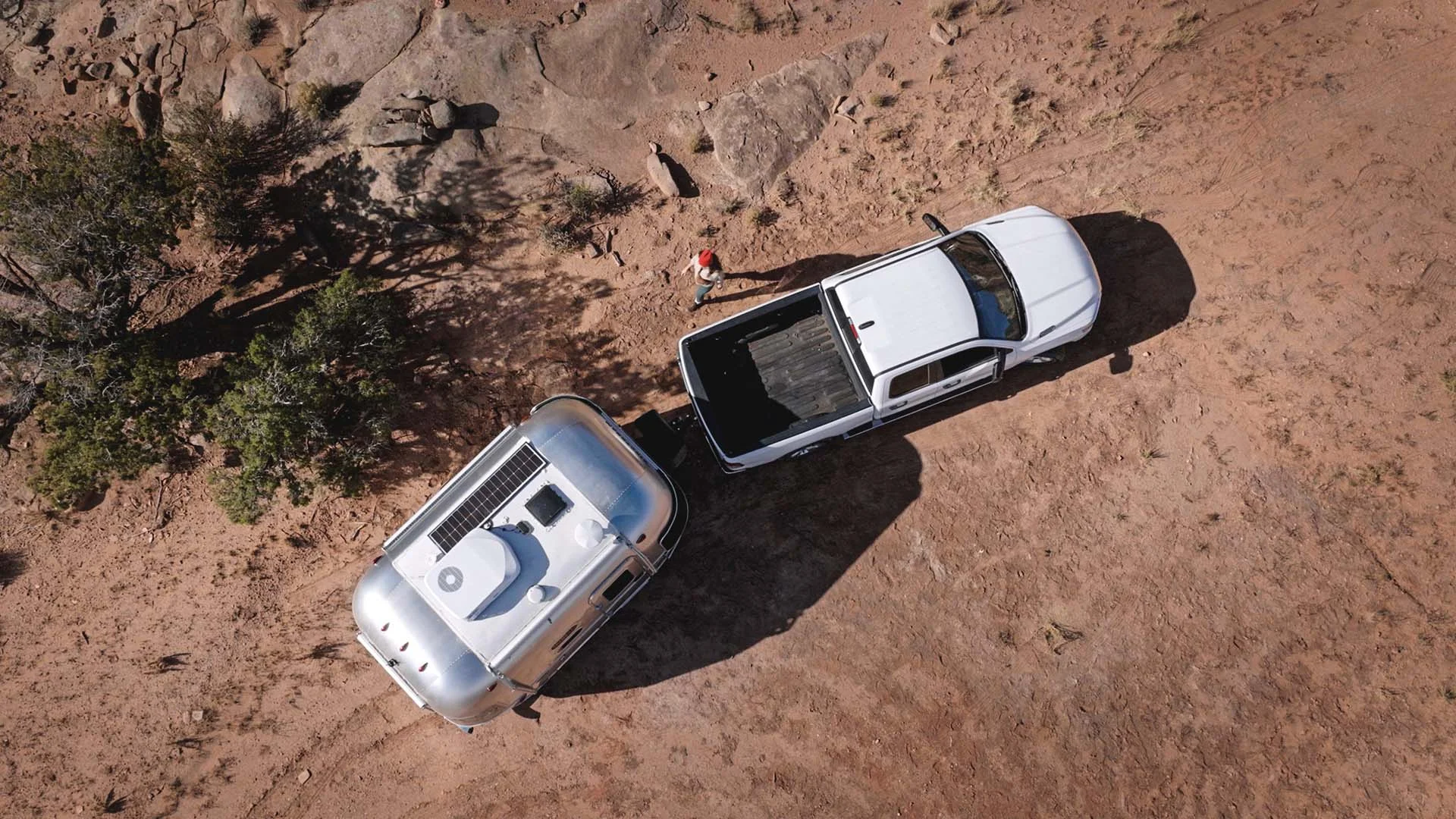 Aerial view of a white pickup truck towing a silver camper trailer on a dirt path, surrounded by scattered rocks and sparse vegetation.