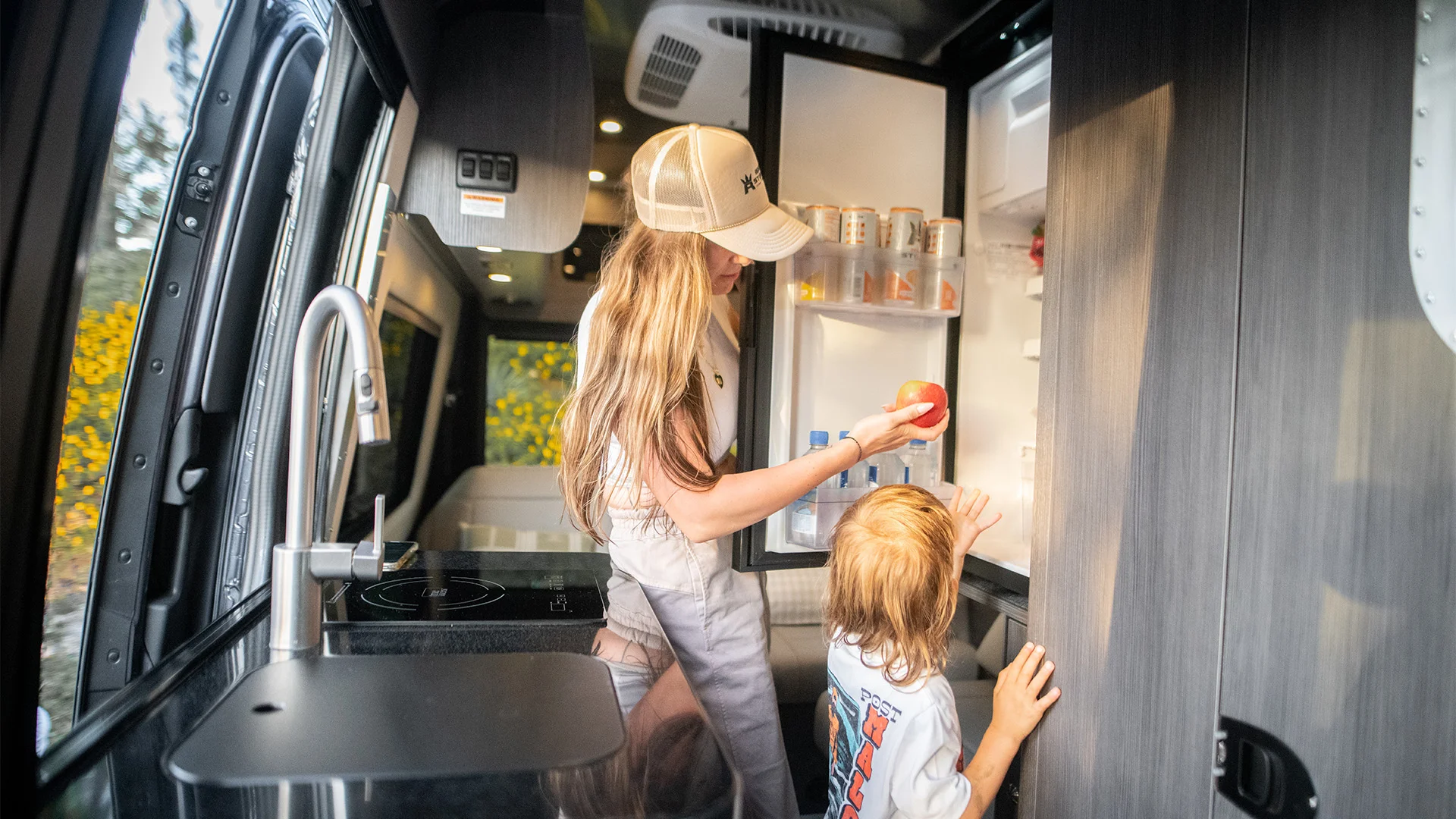 Woman and child inside a camper van kitchen, the woman holding an apple by an open fridge, with sunlight coming through the window.