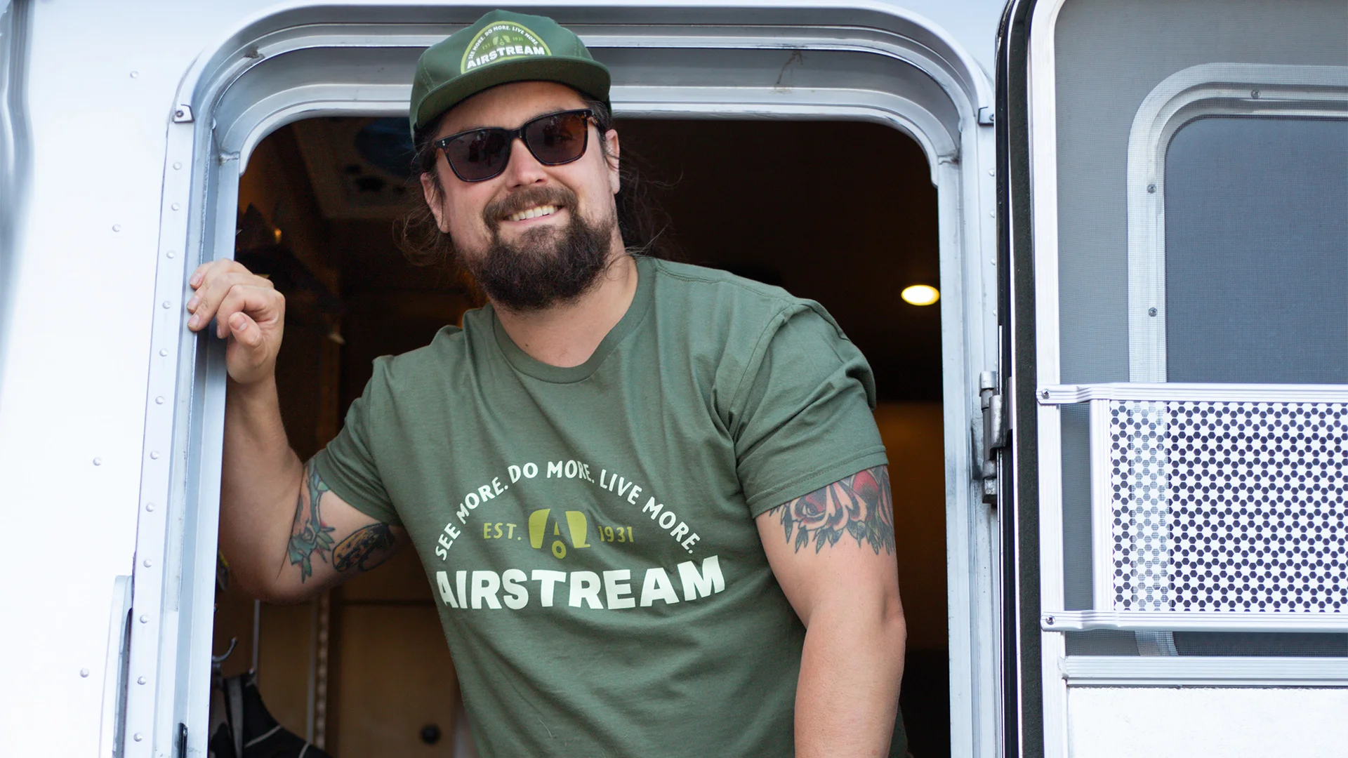 Smiling man with sunglasses and cap, in a green T-shirt with "Airstream" logo, stands in a trailer doorway, revealing arm tattoos.