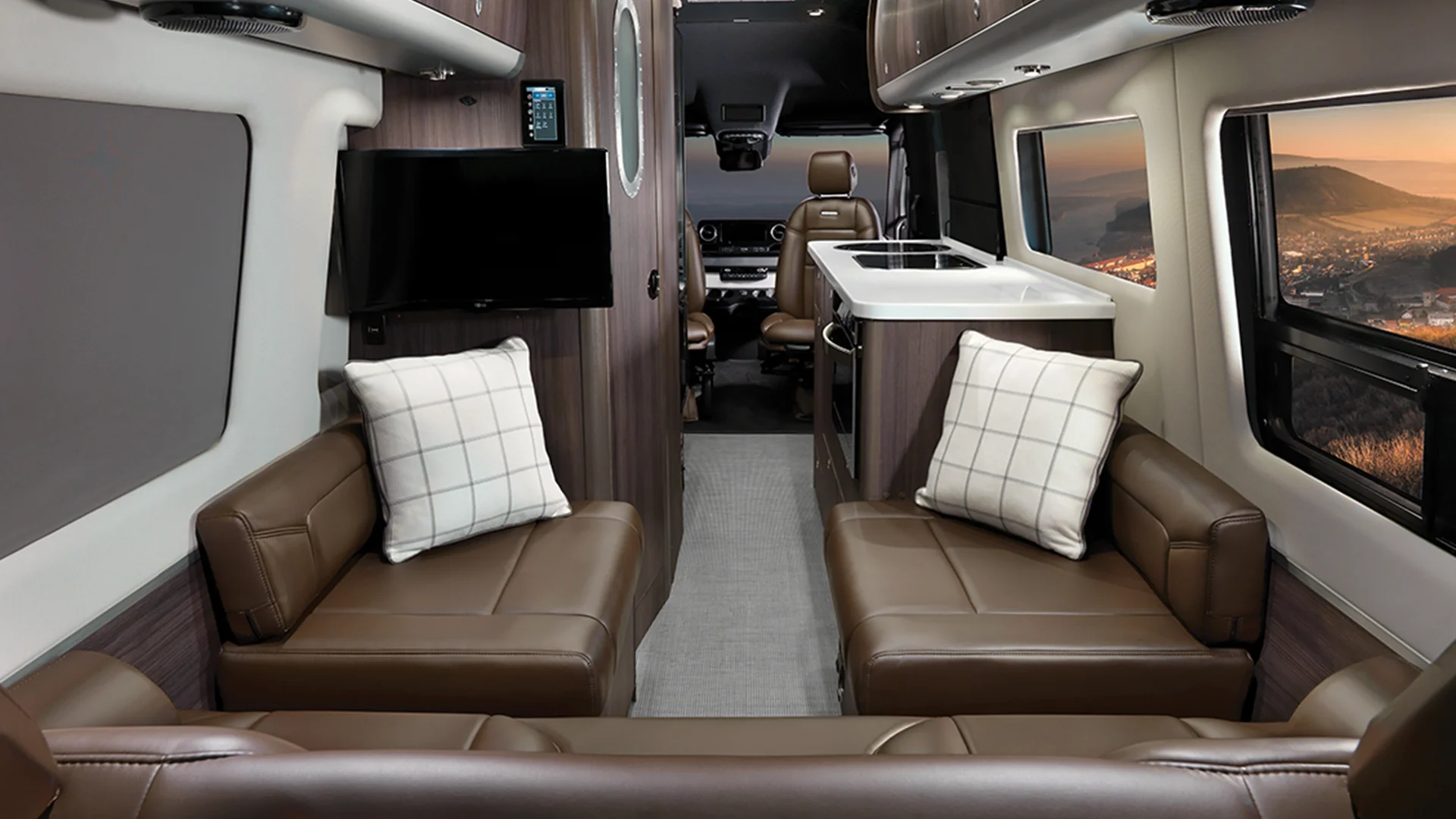 Luxurious RV interior with brown leather seating, plaid pillows, a kitchenette, TV, and large windows showing a scenic sunset view.