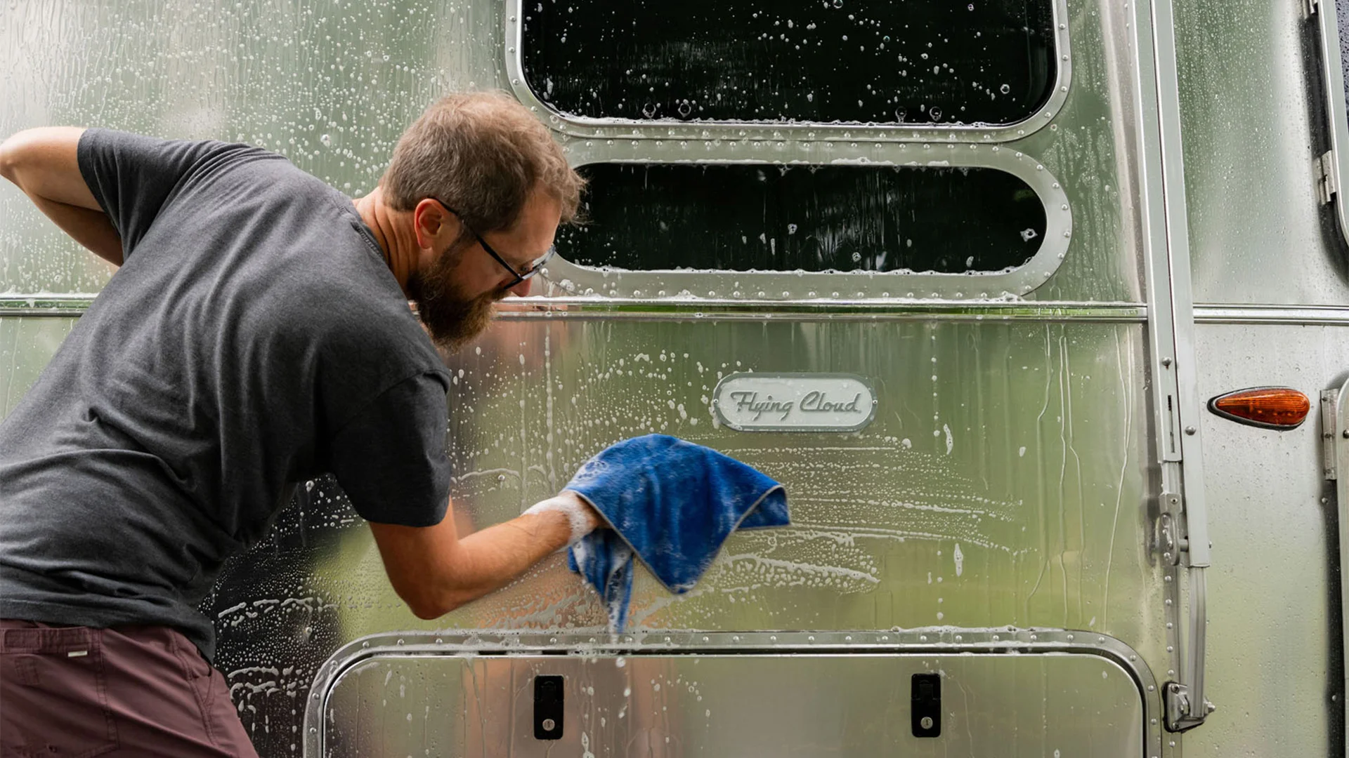 Man cleaning an Airstream trailer with a blue cloth, covered in suds, on a sunny day.