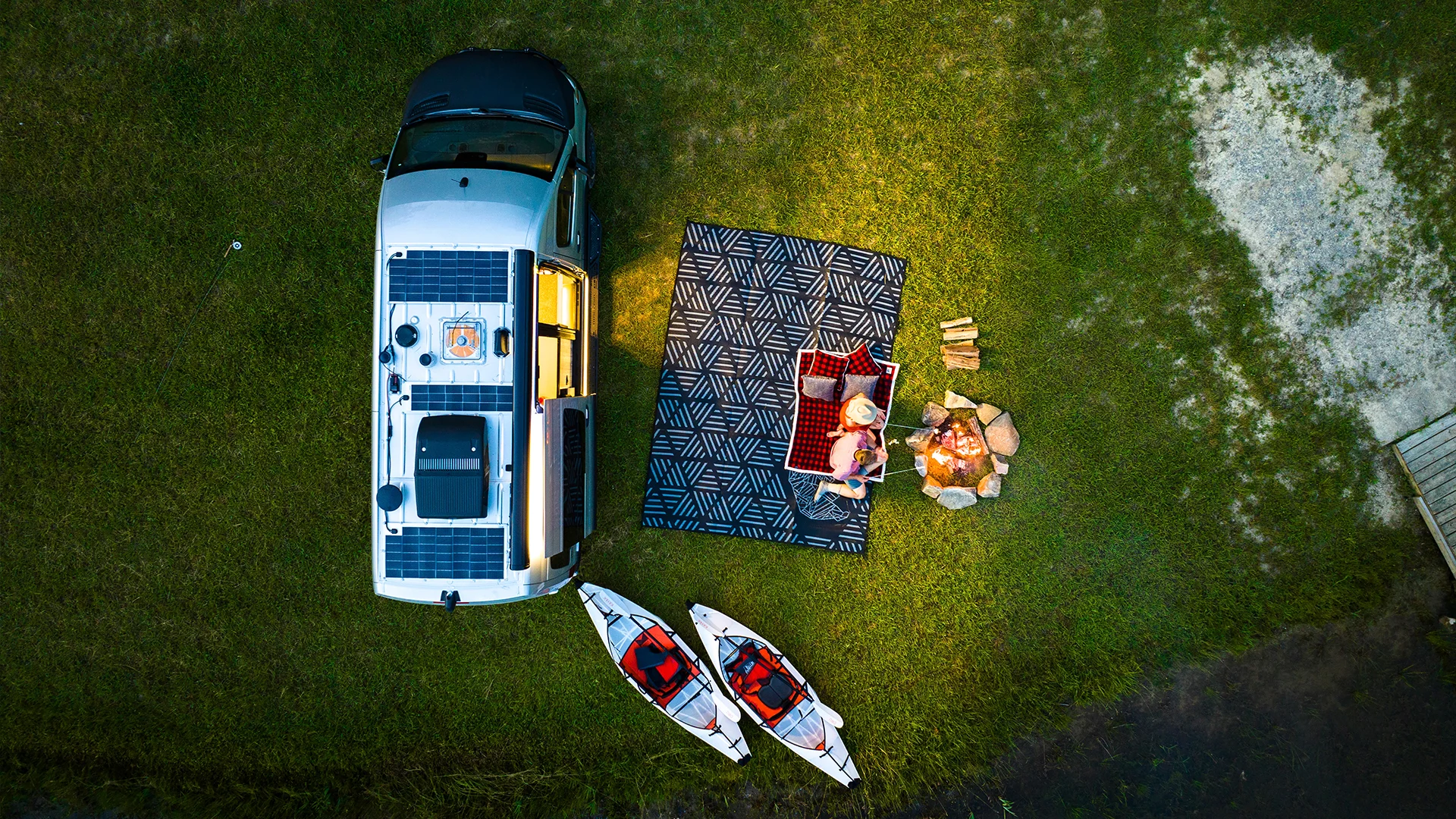 Aerial view of a camper van near a patterned picnic blanket with two people, logs, a campfire, and two kayaks on grassy terrain.