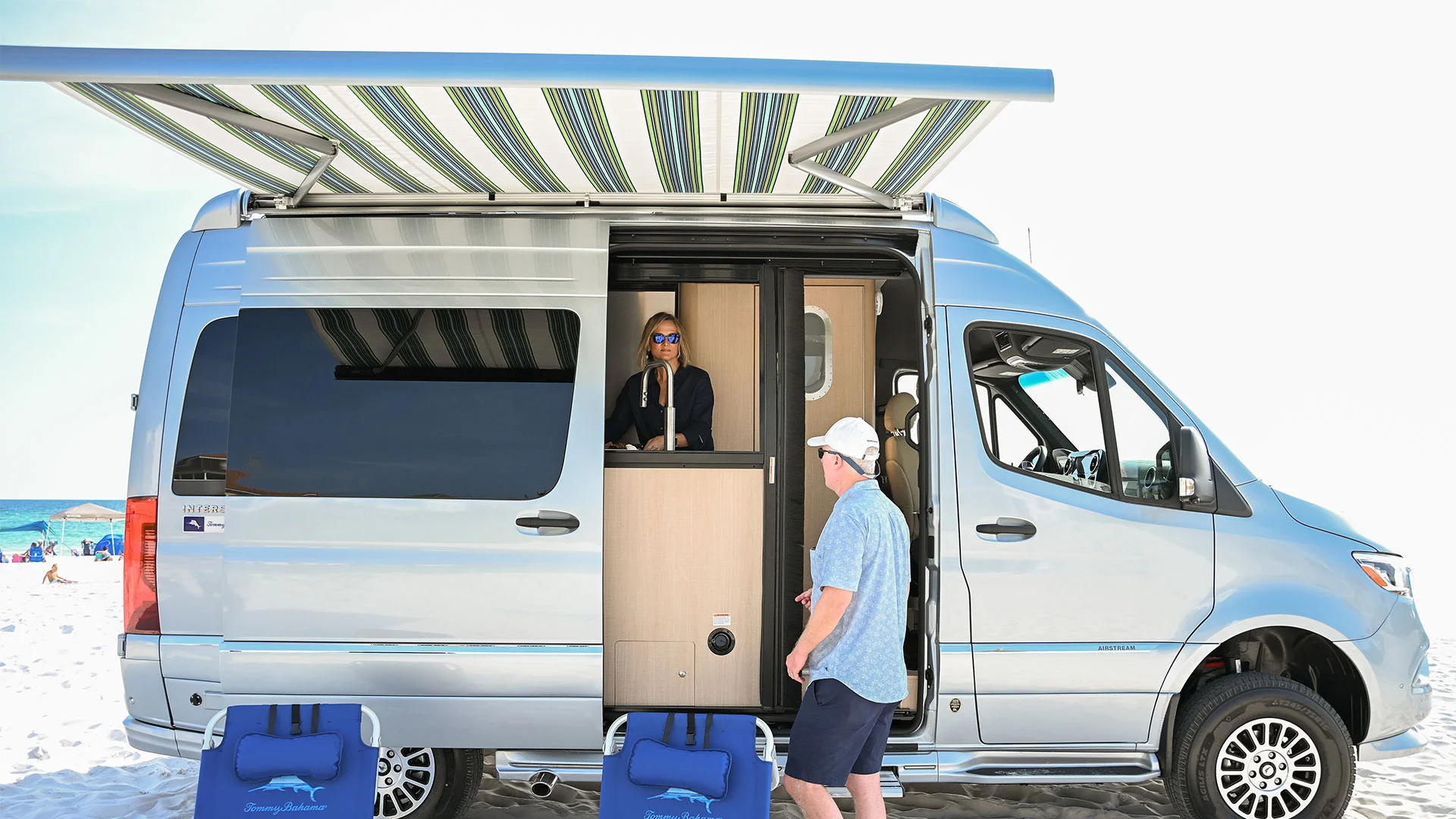 A person stands in a silver camper with a striped awning on a beach, while another approaches. Blue beach chairs are in the foreground.