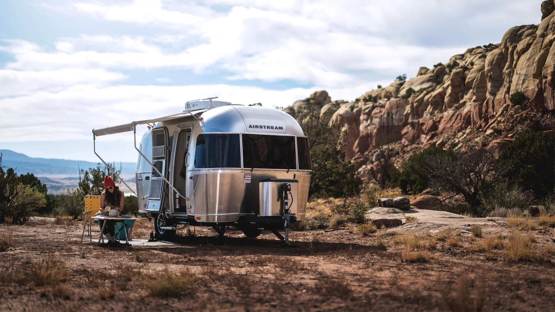 A shiny silver Airstream trailer is parked on dry, rocky terrain near a table where a person is preparing food. Rugged cliffs and sparse vegetation are visible in the background under a partly cloudy sky.