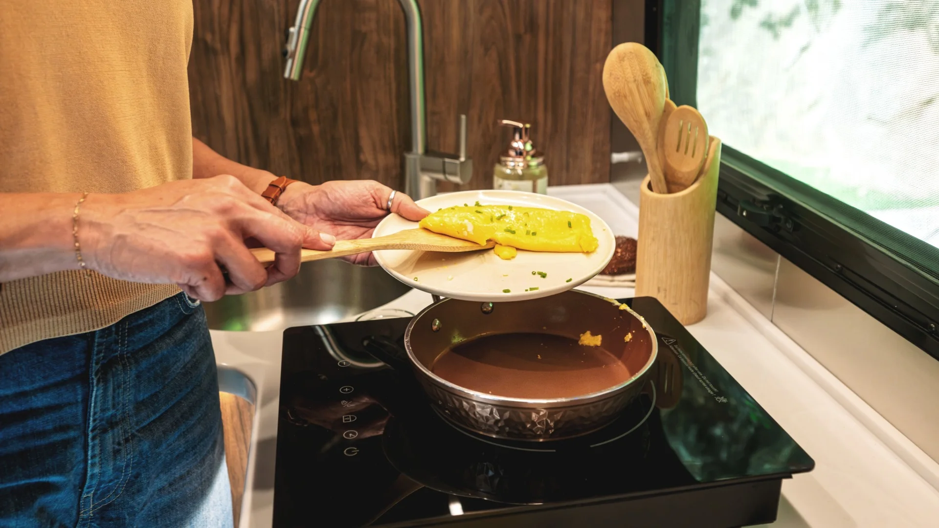 A person transfers a folded omelet with chives from a plate to a pan using a wooden spatula in a kitchen setting.