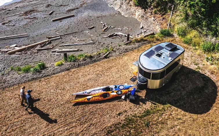 Aerial view of two people walking near a camper trailer parked on grass by a beach, with two kayaks on the ground and driftwood scattered along the shore.
