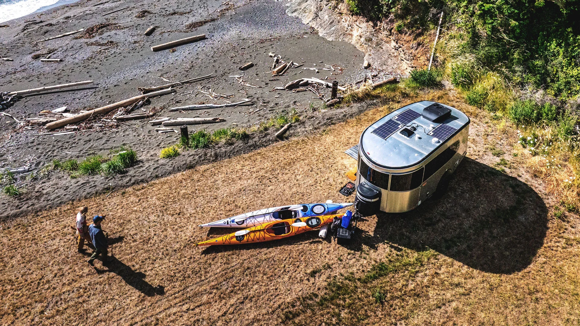 Aerial view of two people walking near a camper trailer parked on grass by a beach, with two kayaks on the ground and driftwood scattered along the shore.