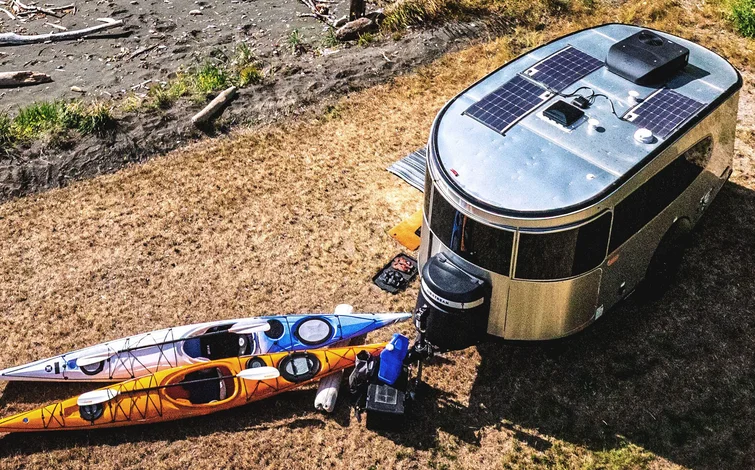 A silver camper van with solar panels is parked on dry grass near a shoreline. Two colorful kayaks, one orange and one blue, are placed beside the camper along with outdoor gear.
