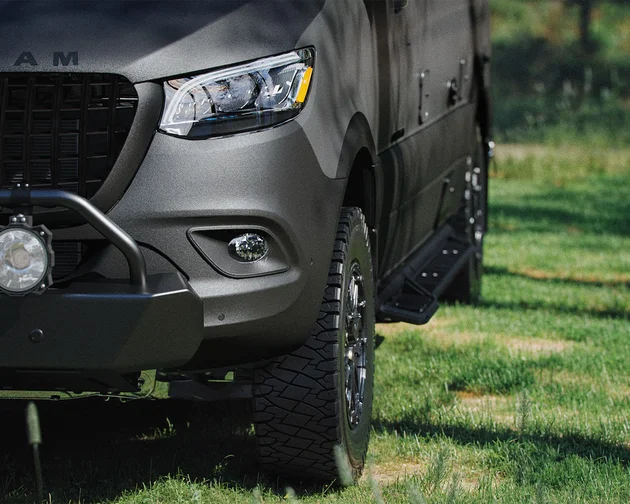 Close-up of a dark gray off-road vehicle with a visible front grille and headlight, parked on grass with trees in the background.